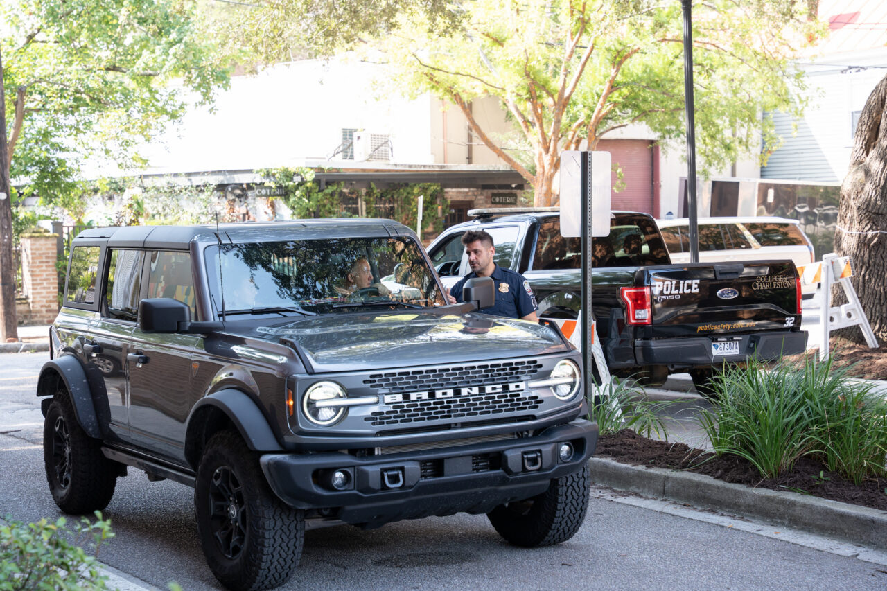 A dark gray Ford Bronco SUV is parked on a street near a police vehicle with officers present.
