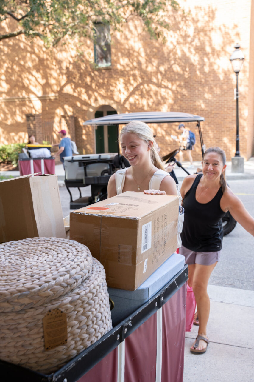 Two individuals smile while pushing a cart loaded with boxes and a woven basket down a sunlit street lined with brick buildings.