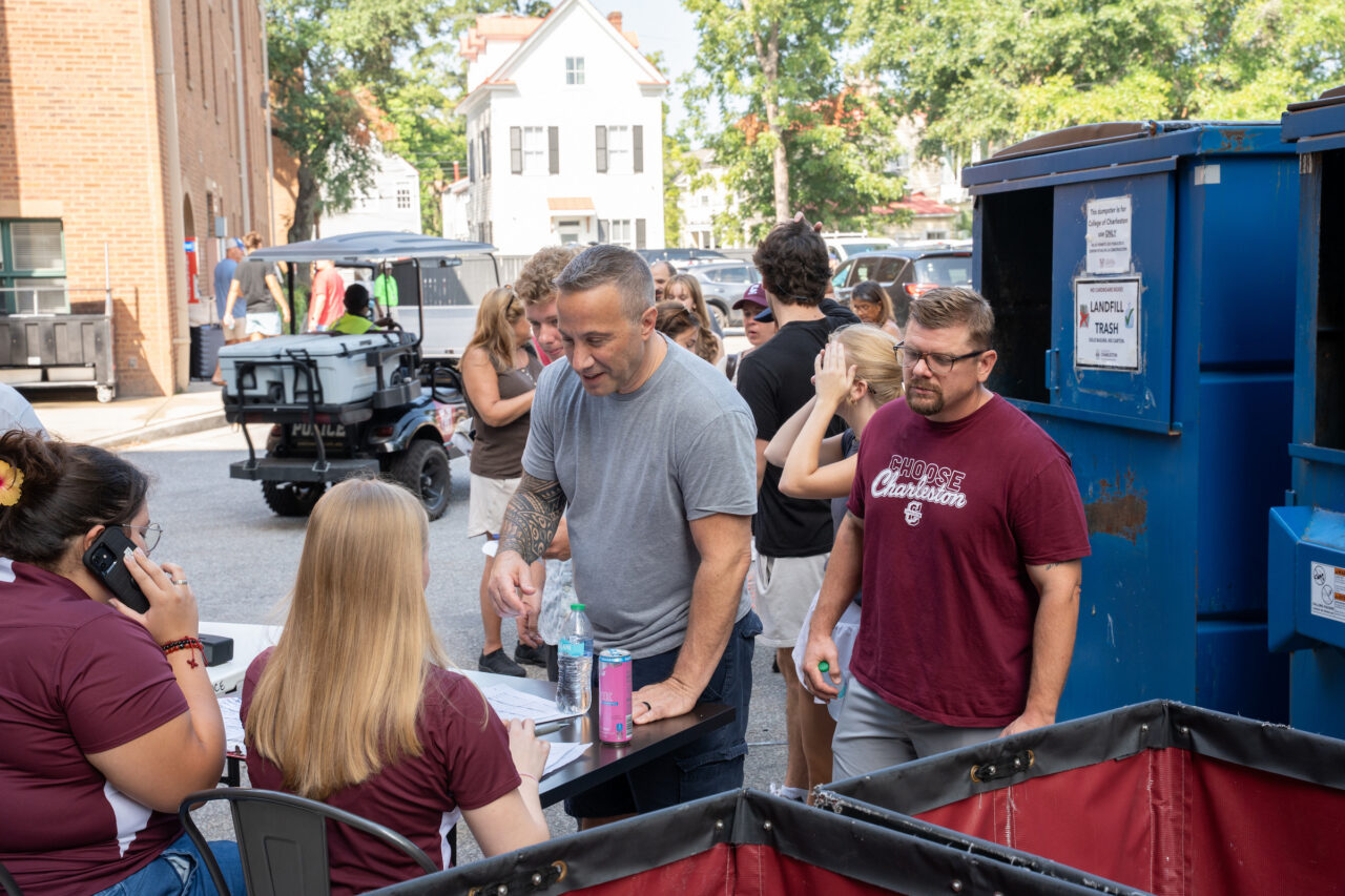 A group of people gather around a table outdoors, with some engaged in conversation and others attending to tasks, near large blue dumpsters and a utility vehicle.