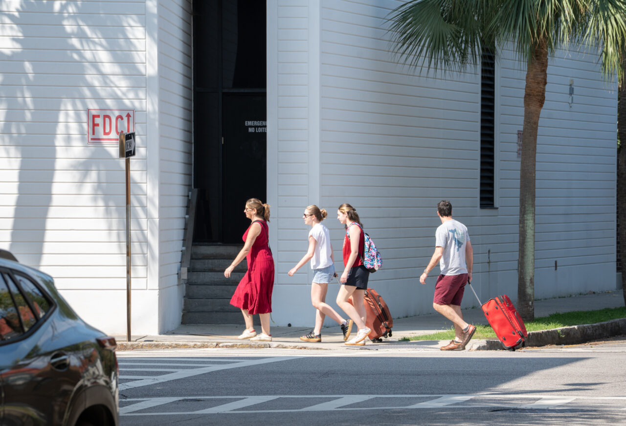 Four individuals walk across a street, with two pulling red suitcases, past a white building marked "FDCT" and a palm tree.