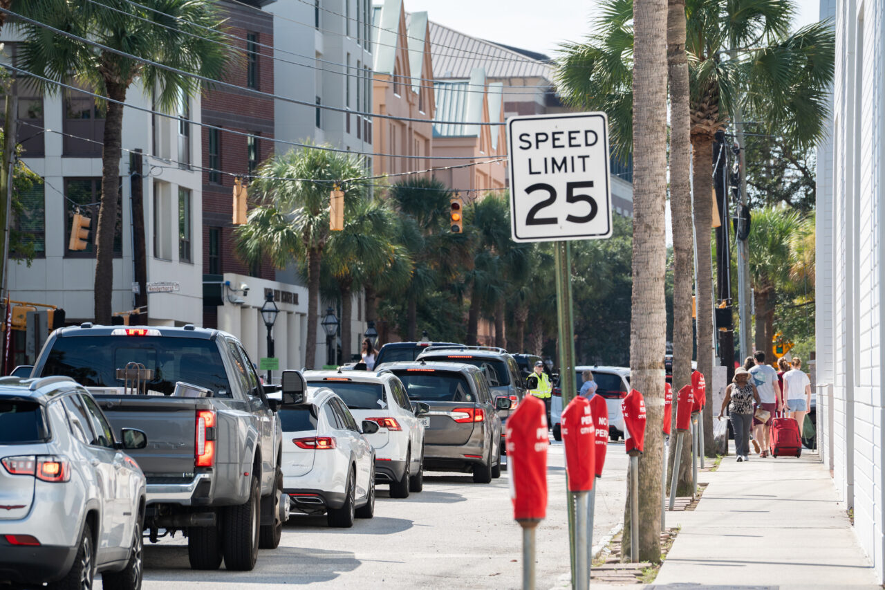 A street lined with palm trees, parked cars, and pedestrians walks past a speed limit sign indicating 25 mph.