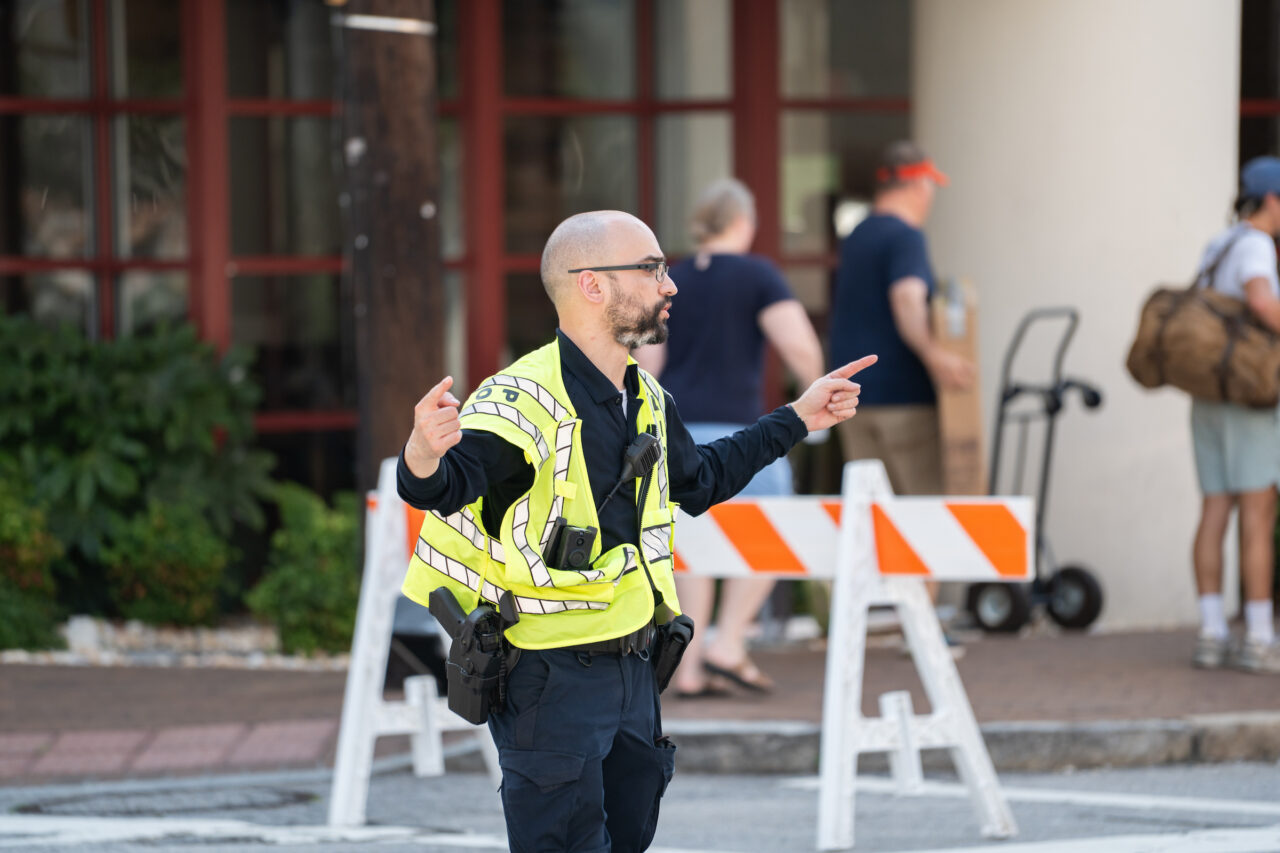 A uniformed officer in a high-visibility vest directs traffic with hand signals near construction barriers.