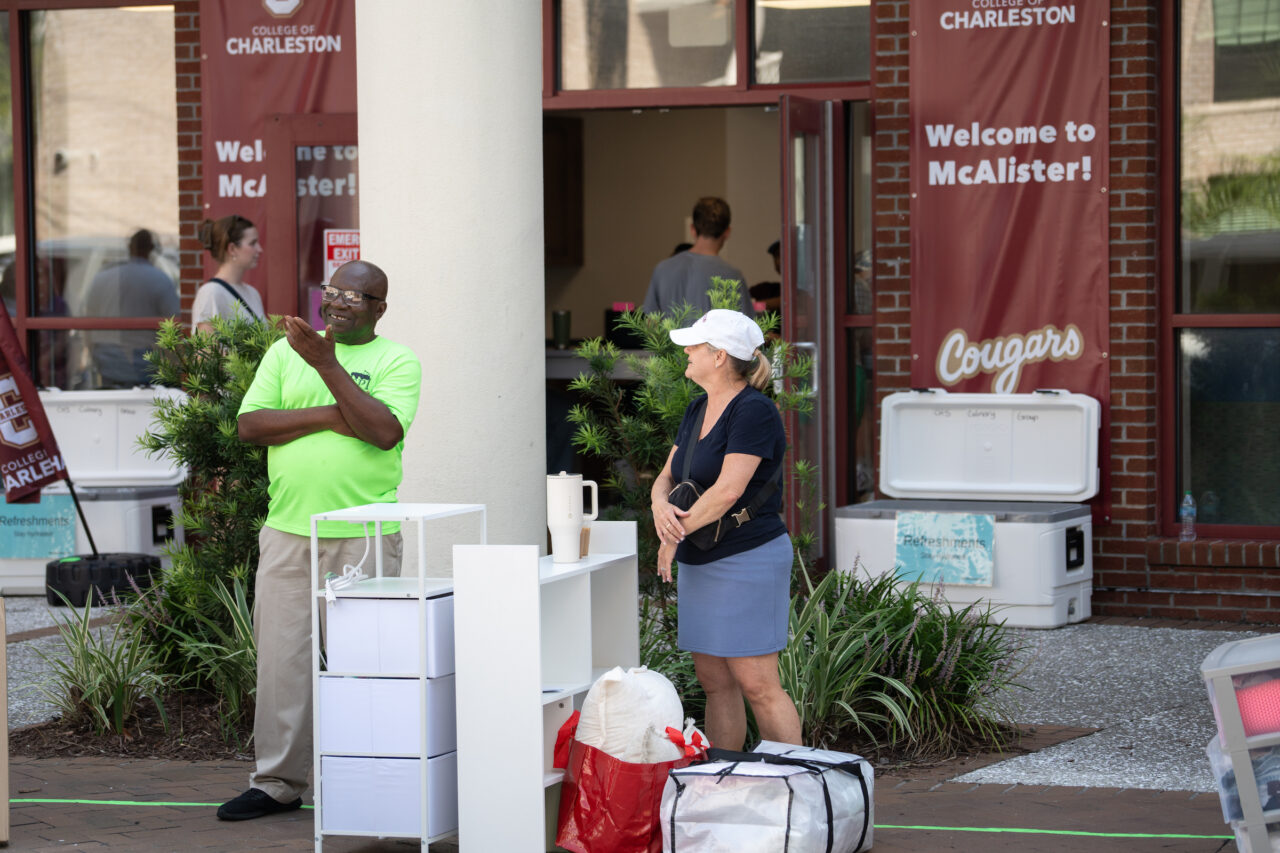 Two individuals stand outside a building with banners welcoming students to the College of Charleston and McAlister, surrounded by coolers and display units.