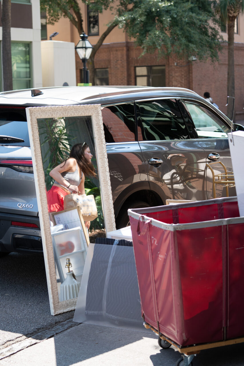 A large ornate mirror leans against a gray SUV parked on a street, next to a red moving cart and various household items.