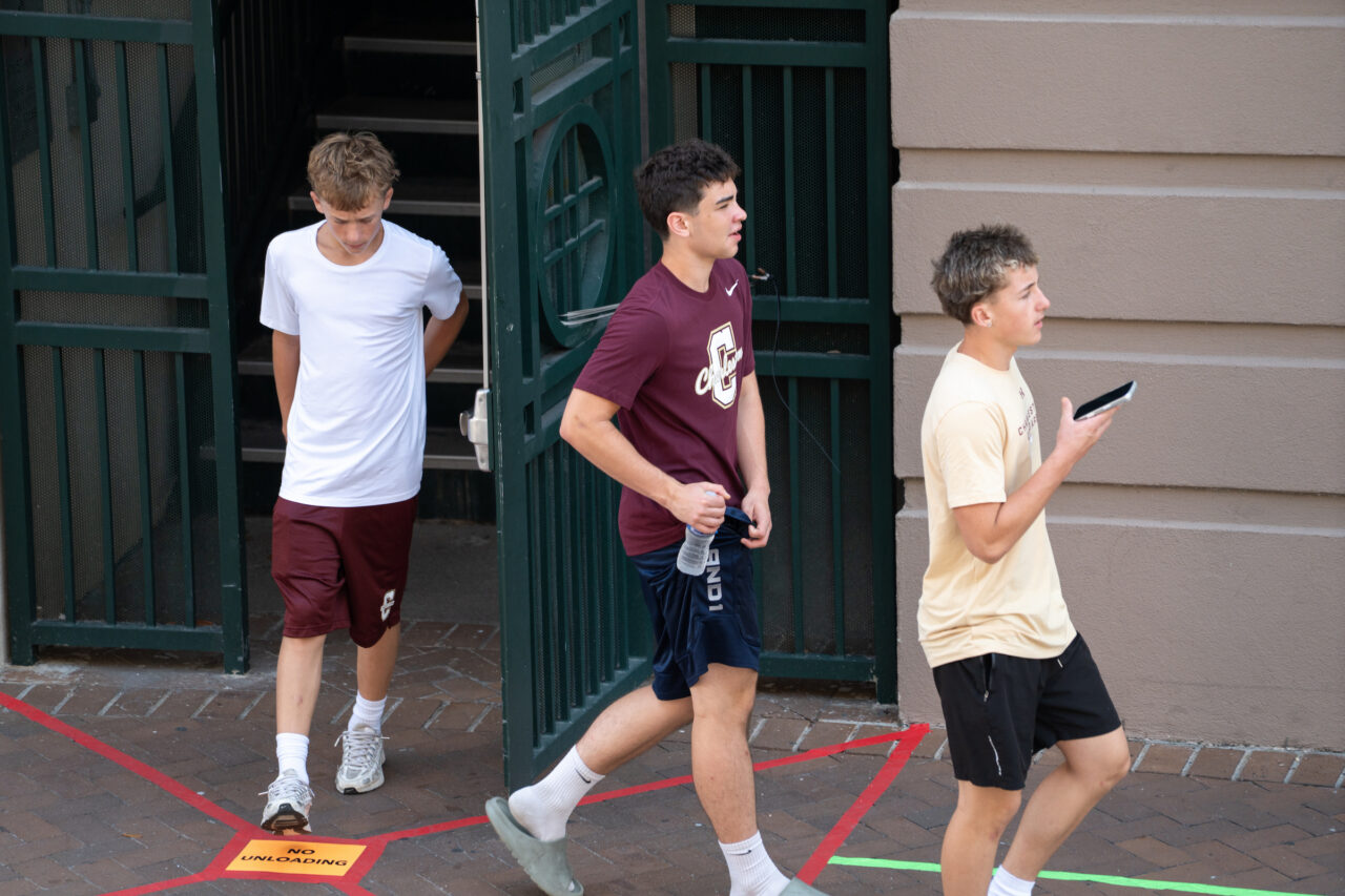 Three young individuals walk out of a building through a green metal gate, with one holding a phone and another carrying a water bottle.