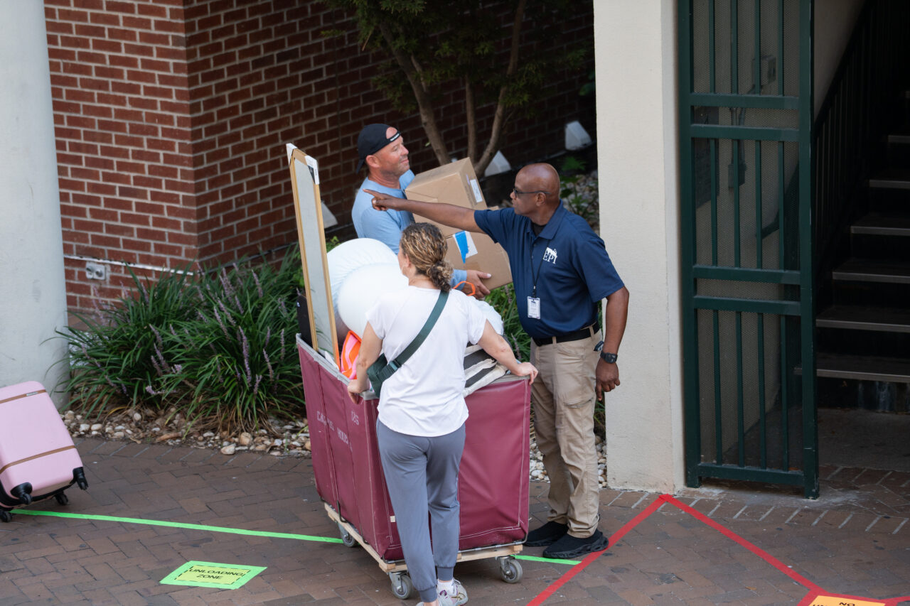 Three individuals collaborate to load items onto a cart outside a building, with one person gesturing toward a box.