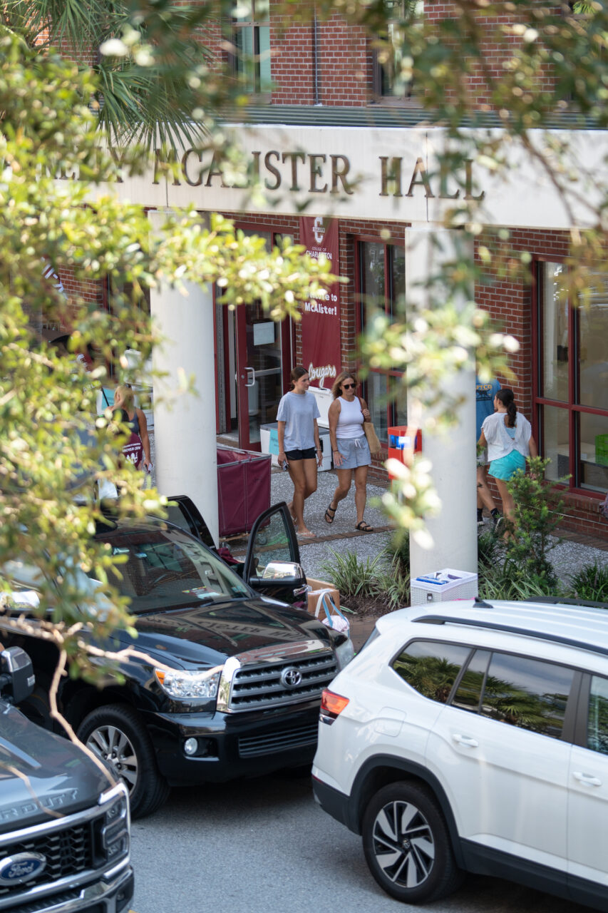 People walk near a building labeled "McAlister Hall" as vehicles are parked nearby, with tree branches partially obscuring the view.