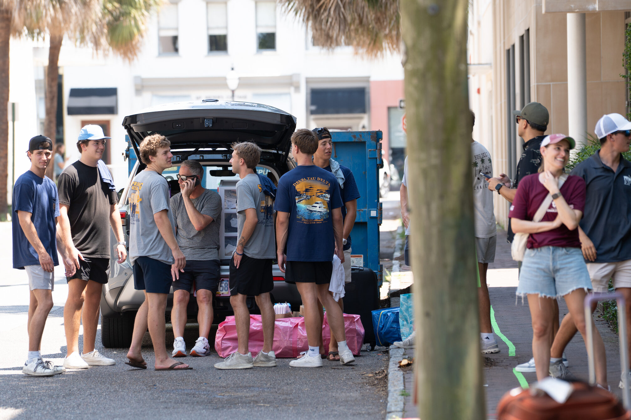 A group of young people gather around an open car trunk on a sunny street lined with palm trees, while others walk by on the sidewalk.
