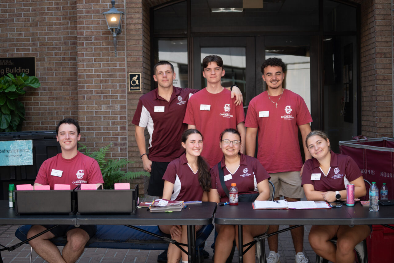 A group of seven students in matching maroon shirts pose outdoors at a table with laptops and supplies, likely for an event or campus activity.