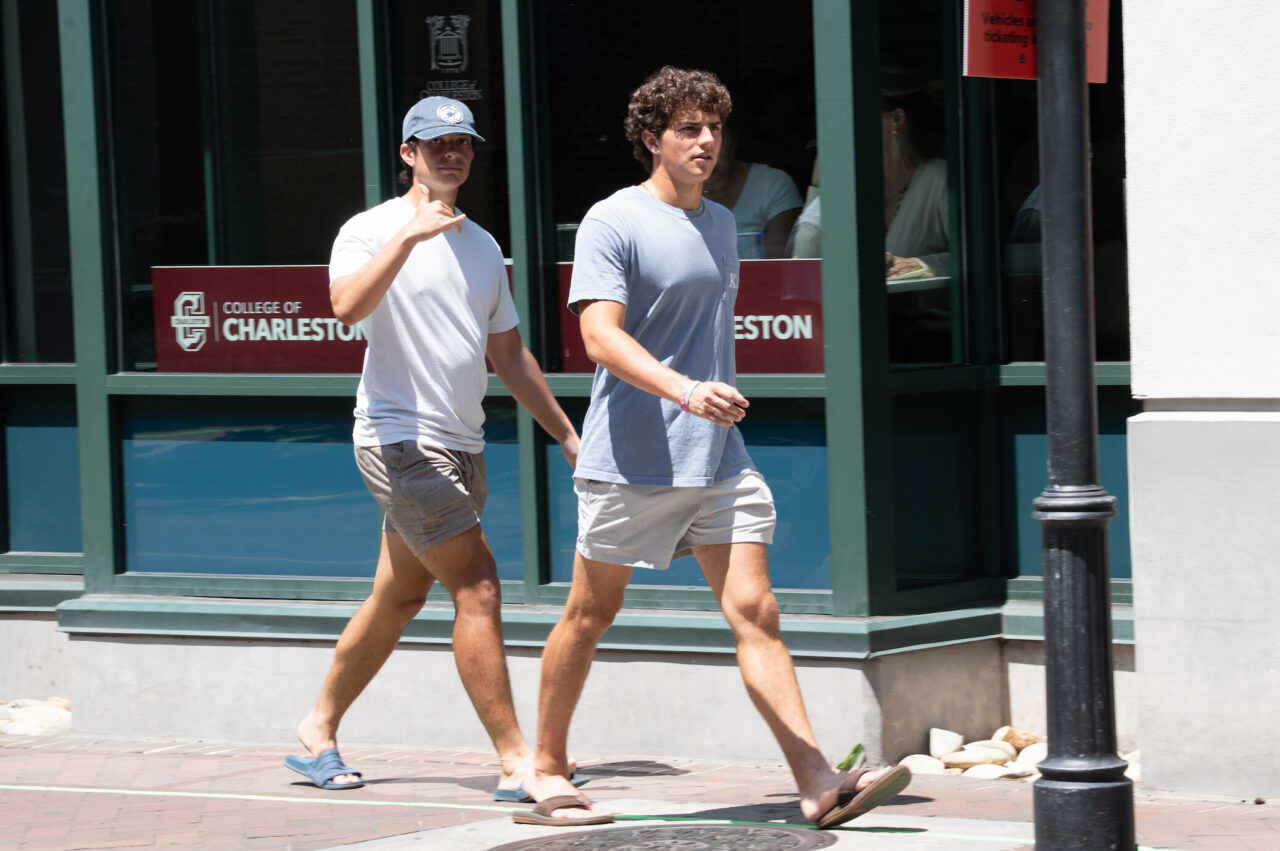 Two young people walk side by side on a sunny sidewalk in front of a building with "College of Charleston" signage.