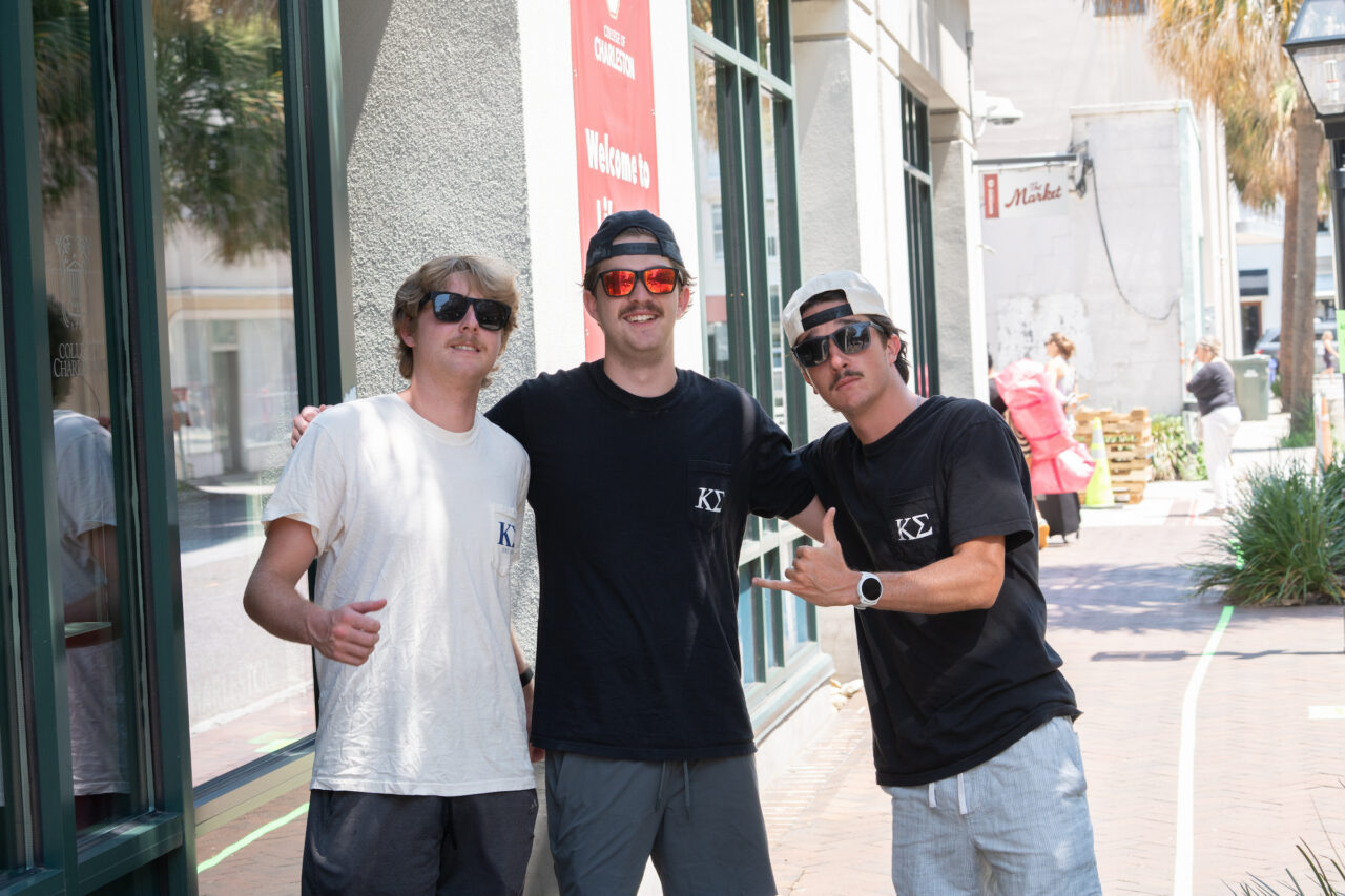 Three individuals pose together outdoors on a sunny day, wearing casual clothing and sunglasses, with one giving a thumbs-up and another making a hand gesture.