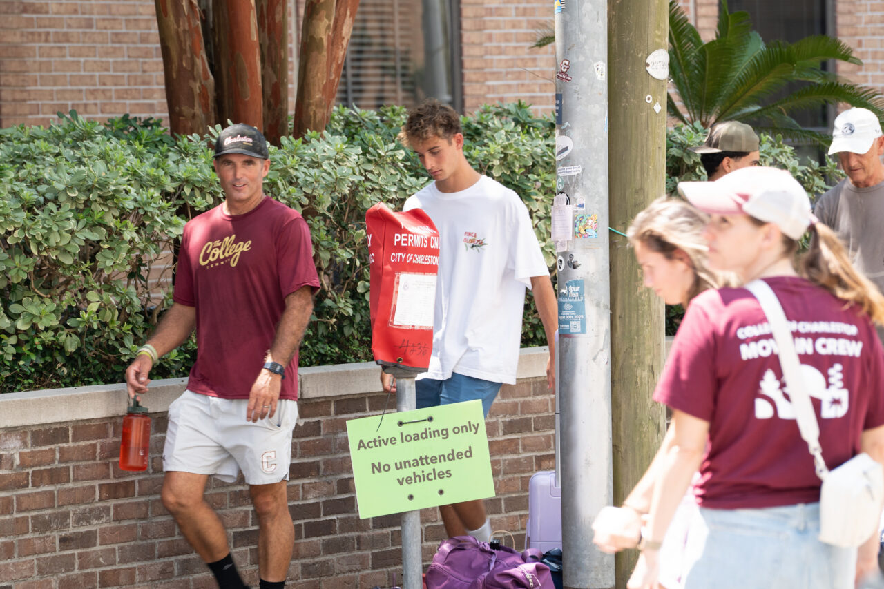 A group of people, including a coach and athletes, stand near a parking meter and sign on a brick-lined street.