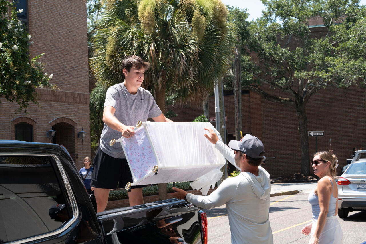 Two individuals carefully lift a large, decorated object from the bed of a black pickup truck on a sunny street.