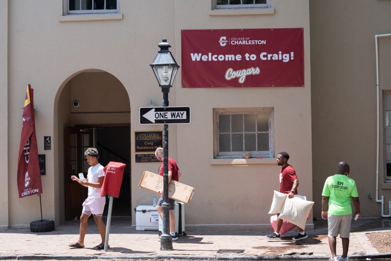 Students and staff carry boxes and pillows outside Craig Hall at the College of Charleston, with a "Welcome to Craig!" banner displayed above the entrance.