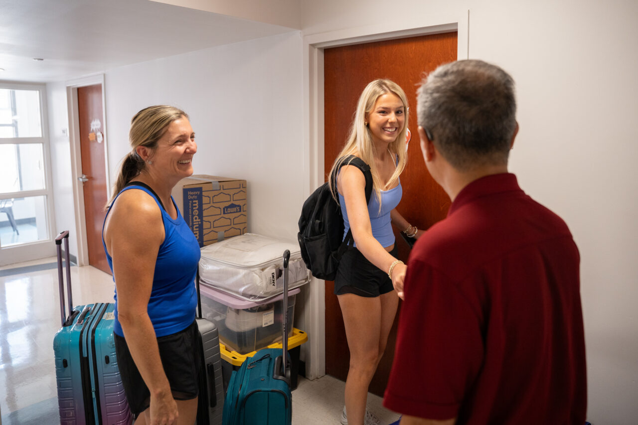A young person smiles while shaking hands with an adult in a hallway, as another adult stands nearby with luggage and boxes.
