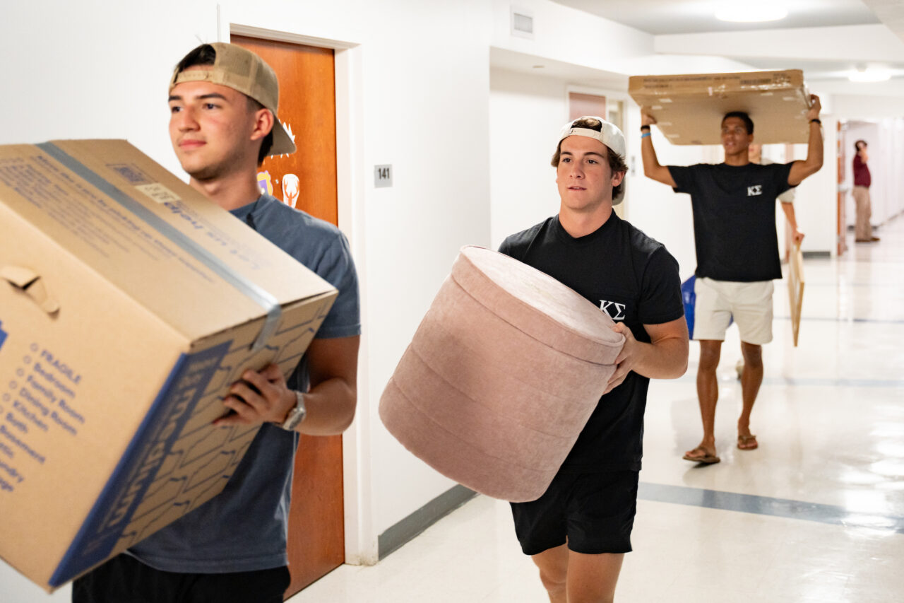 Three young people carry boxes and furniture down a hallway while others follow behind.