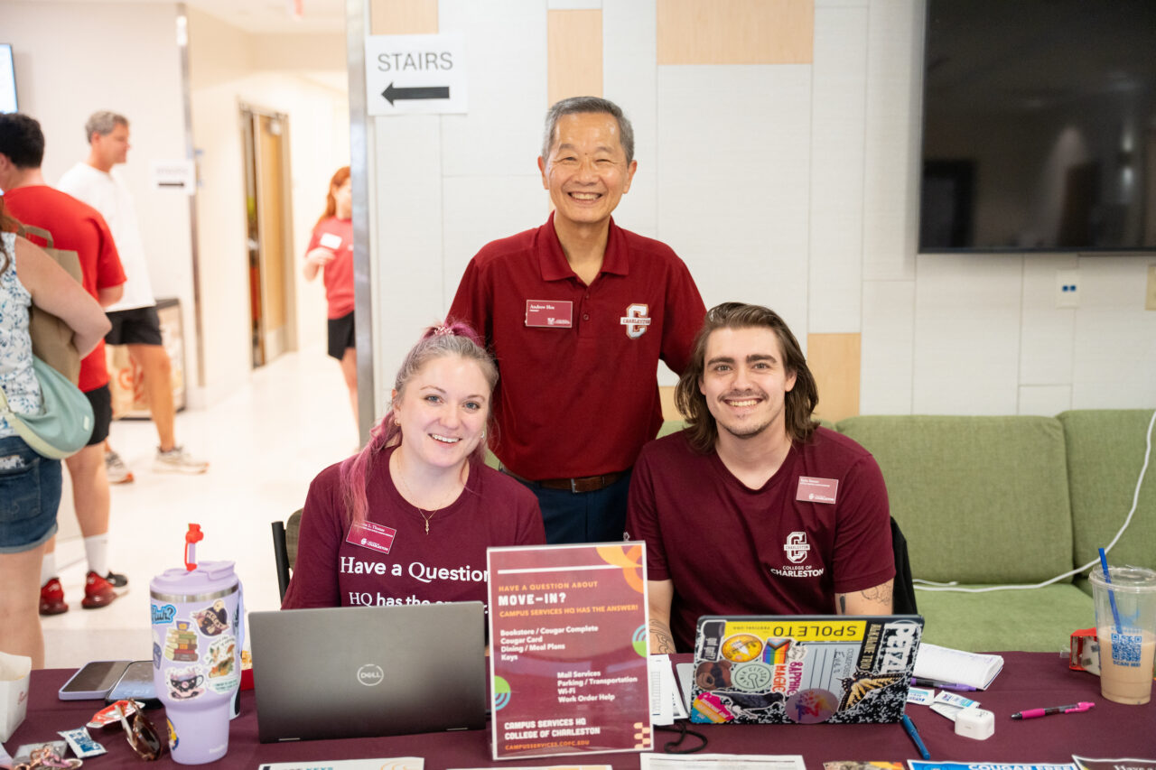 Three individuals in maroon shirts stand behind a table with laptops and informational materials, smiling for the camera at what appears to be a campus event.