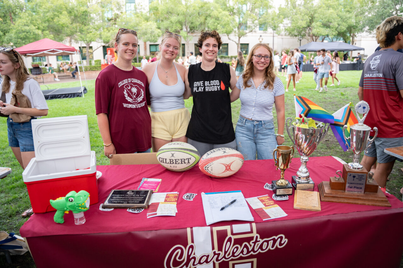 Four individuals stand behind a table covered with a red cloth displaying rugby equipment, trophies, and promotional materials for Charleston.