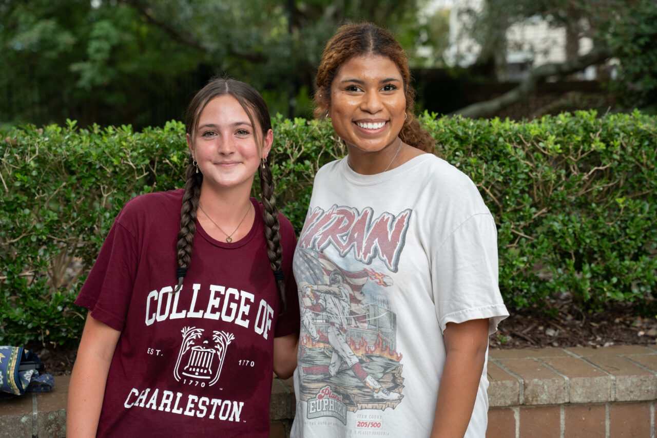 Two young individuals stand together outdoors, smiling, with one wearing a maroon "College of Charleston" shirt and the other in a white graphic tee.