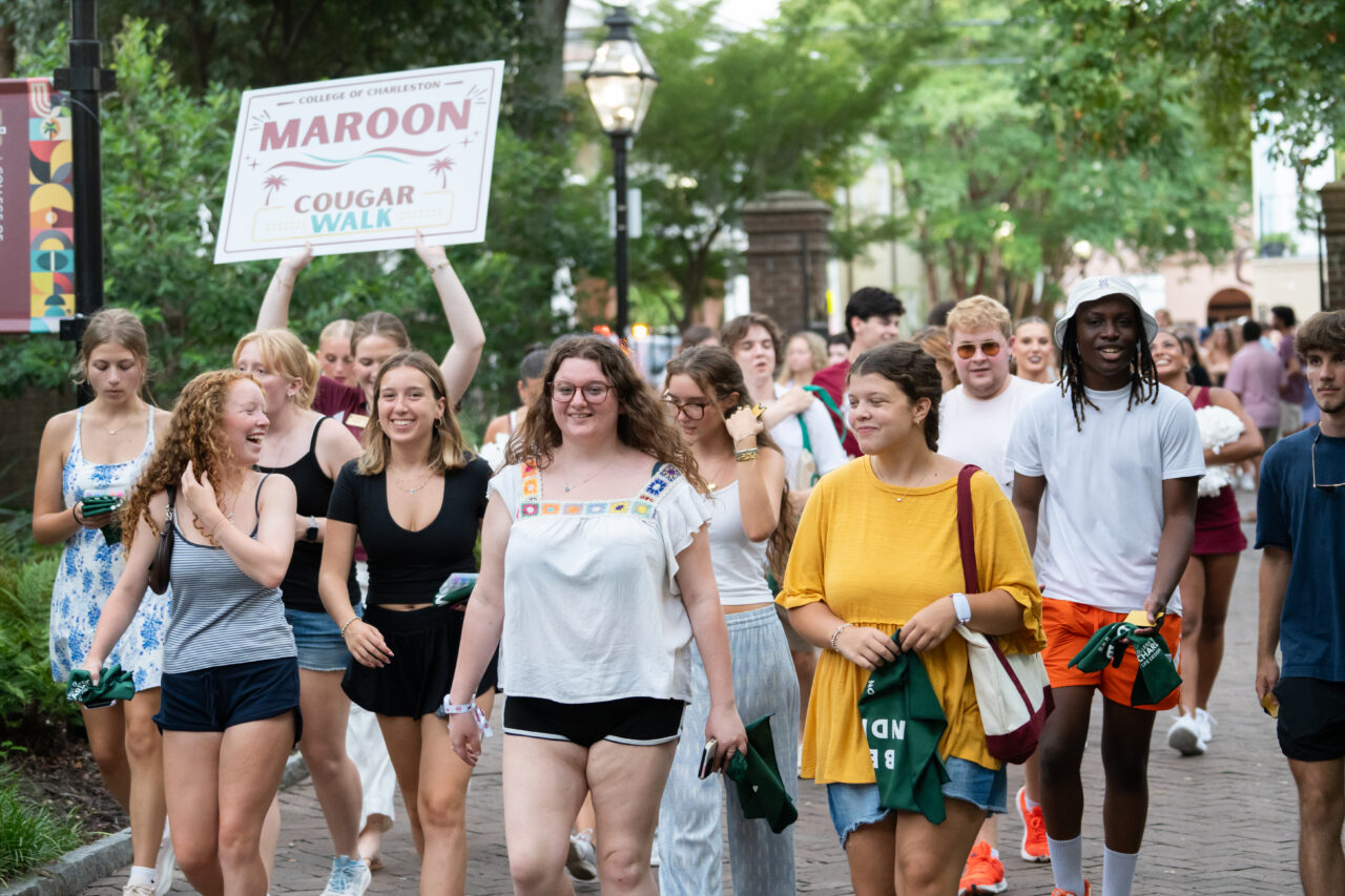 A group of students walk together on a campus pathway, with one person holding a sign that reads "Maroon Cougar Walk."