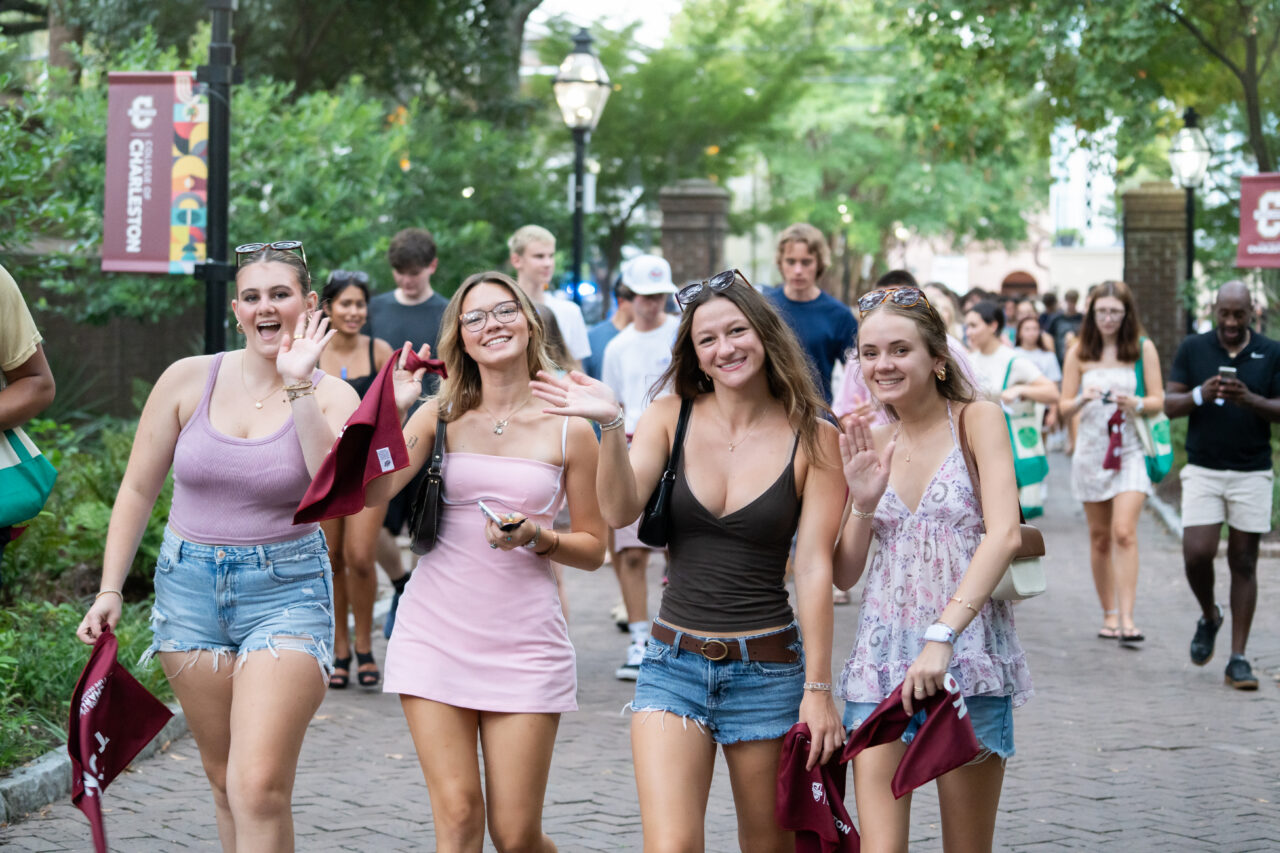 Four young people walk together on a campus pathway, smiling and waving, while others move around them in the background.