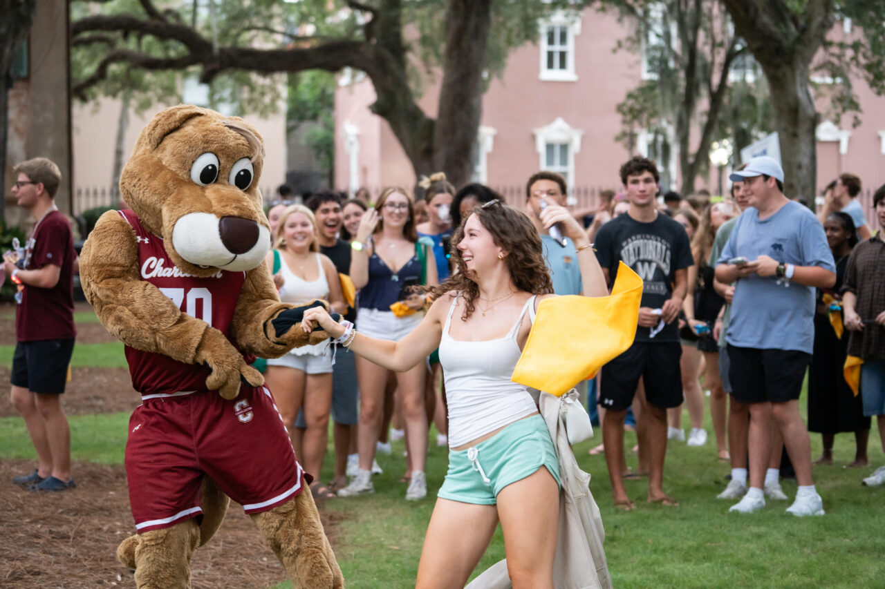 A person in a large brown mascot costume dances with a young individual holding a yellow flag, surrounded by a crowd on a grassy area.
