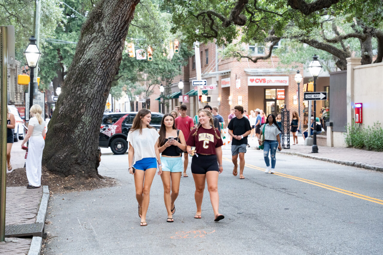 Three young people walk along a tree-lined street in a bustling urban area, with shops and other pedestrians visible in the background.