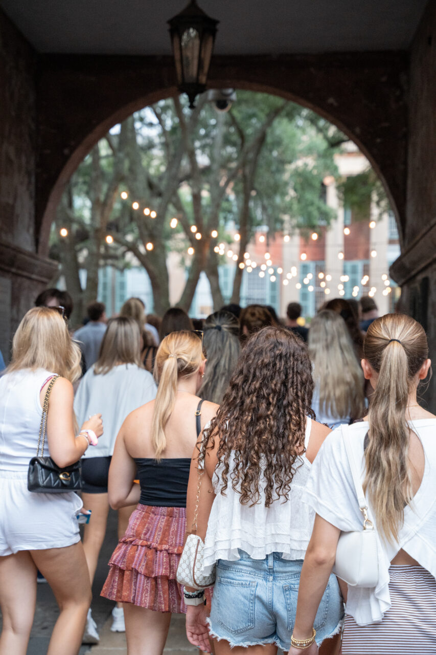 A group of people walk through an arched passageway toward a tree-lined area adorned with string lights.