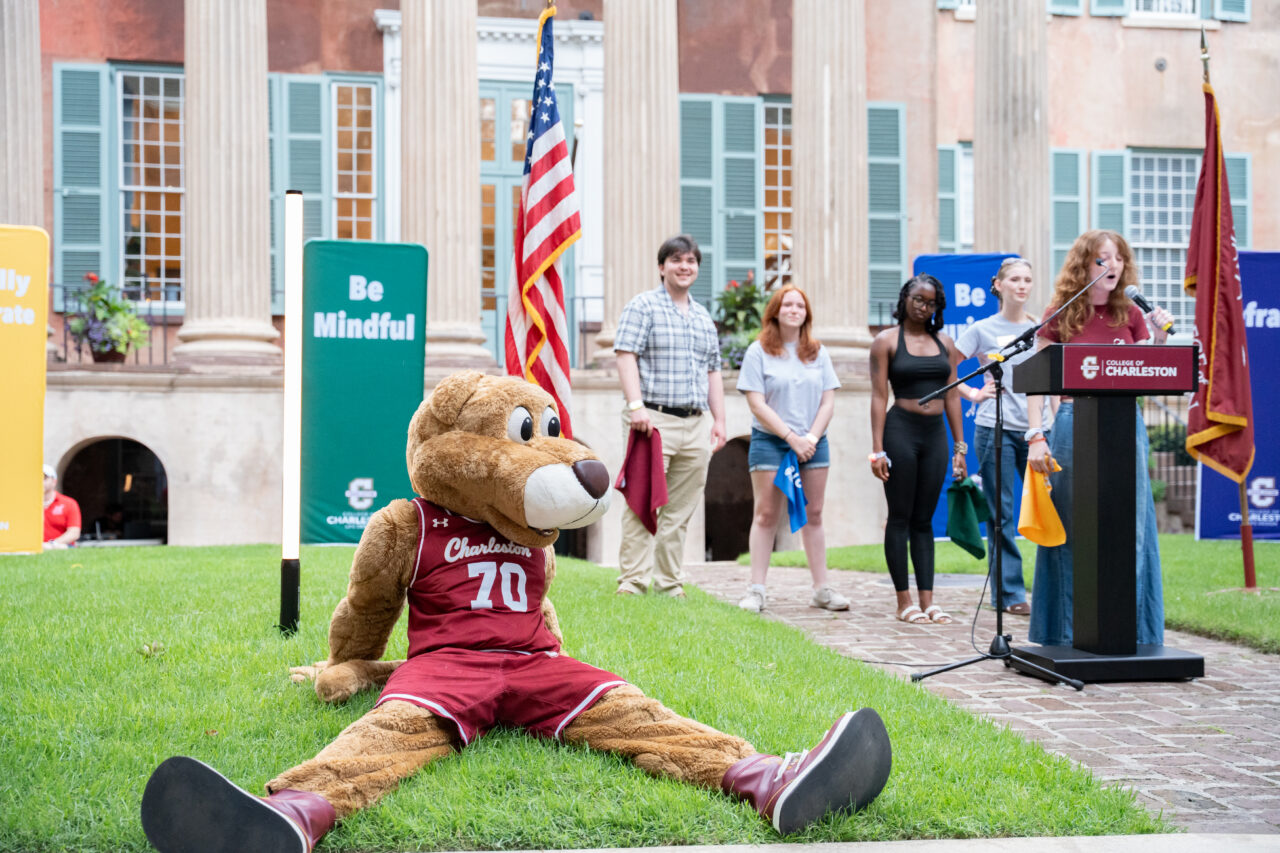 A large mascot in a basketball uniform sits on grass near a podium where a person speaks, with several others standing nearby and flags visible in the background.