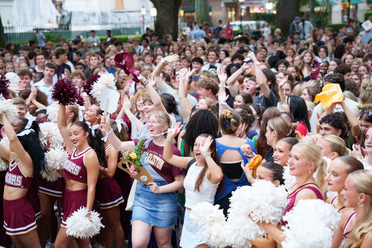 A lively crowd of young people, including cheerleaders, cheerfully wave pom-poms and hats at an outdoor event.