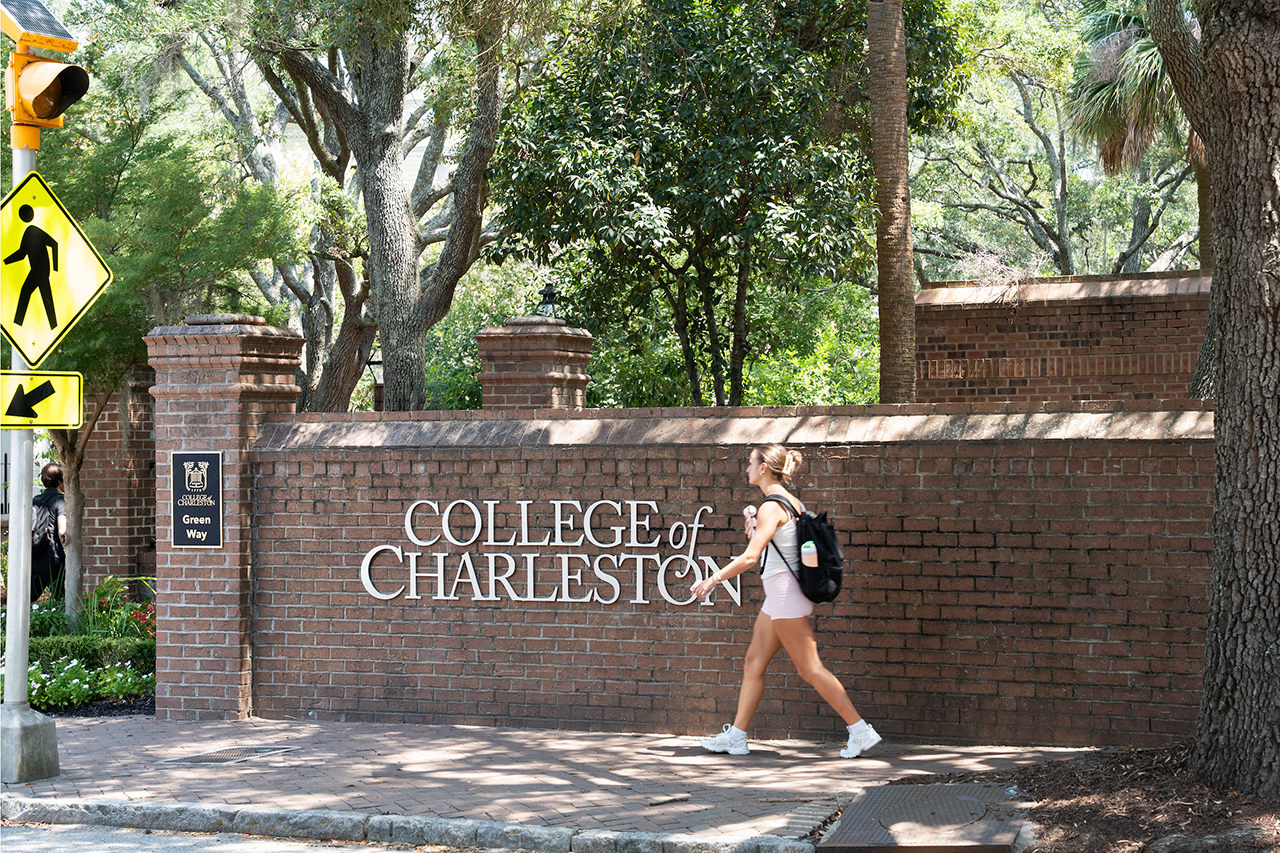 A person walks past a brick wall bearing the sign "College of Charleston" on a sunny day, with trees and traffic signs visible nearby.