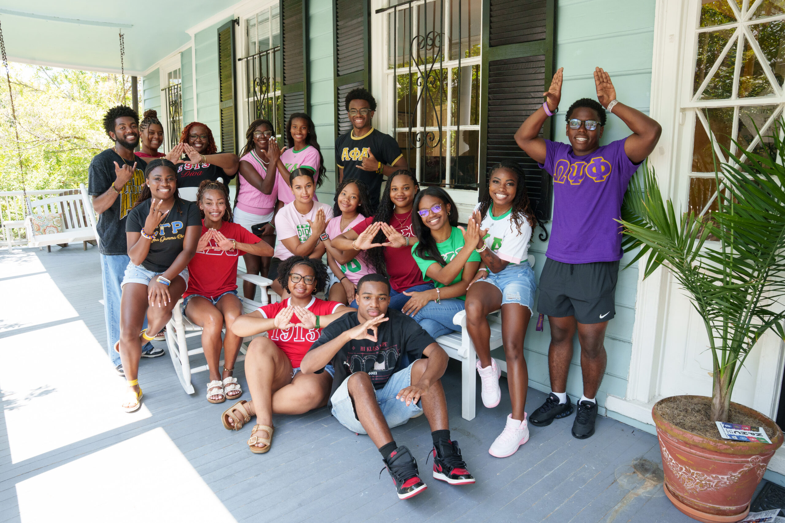 A group of young people wearing fraternity shirts pose cheerfully on a porch, making hand gestures and smiling for the camera.