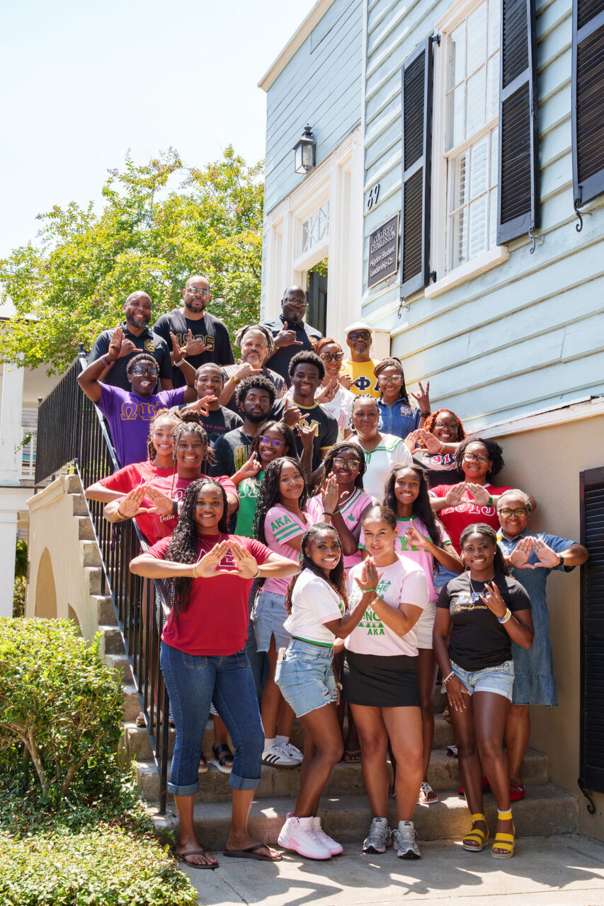 A diverse group of young people and adults pose cheerfully on outdoor steps in front of a historic building, many making heart shapes with their hands.