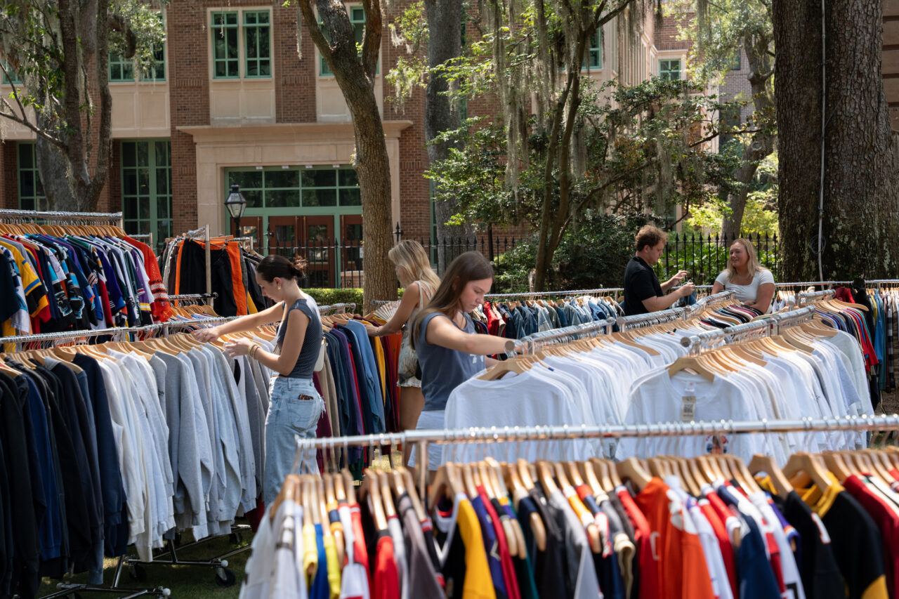 People browse through racks of clothing at an outdoor market under trees.