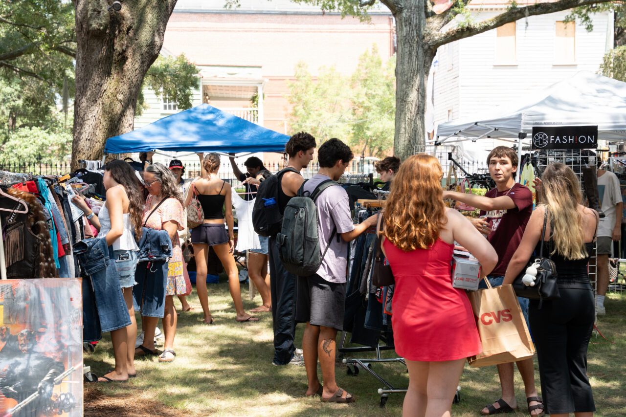 A group of people browse clothing racks at an outdoor market under trees and tents.