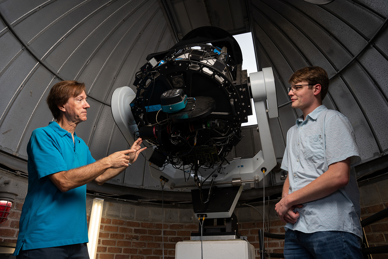 Two individuals stand inside a telescope dome, engaged in discussion while pointing toward a large, complex astronomical instrument.