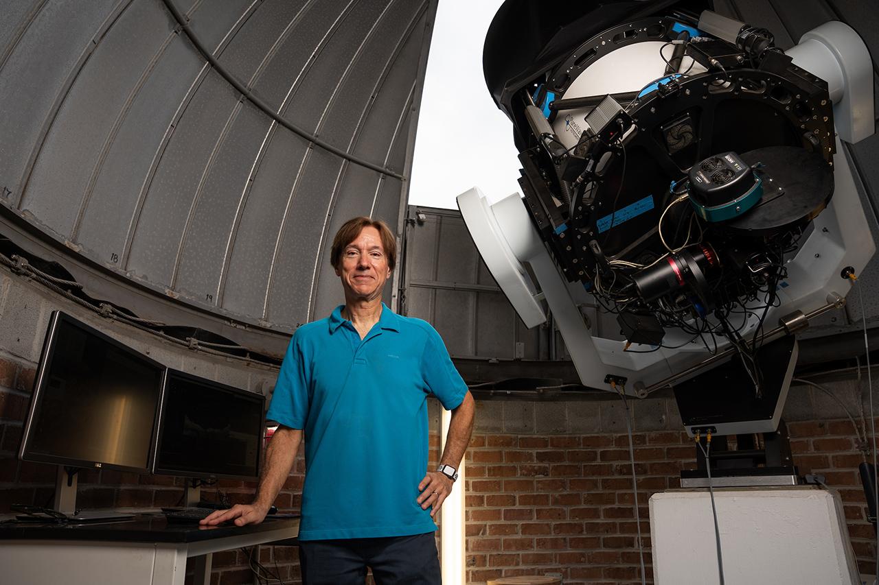 A person stands in an observatory next to a large telescope, with computer monitors on a desk nearby.