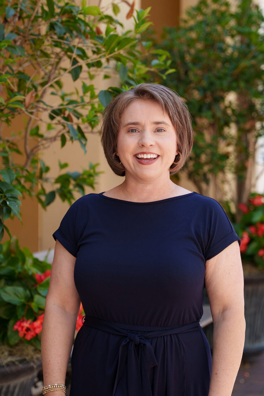 A smiling person in a navy blue dress stands outdoors, surrounded by potted plants and greenery.