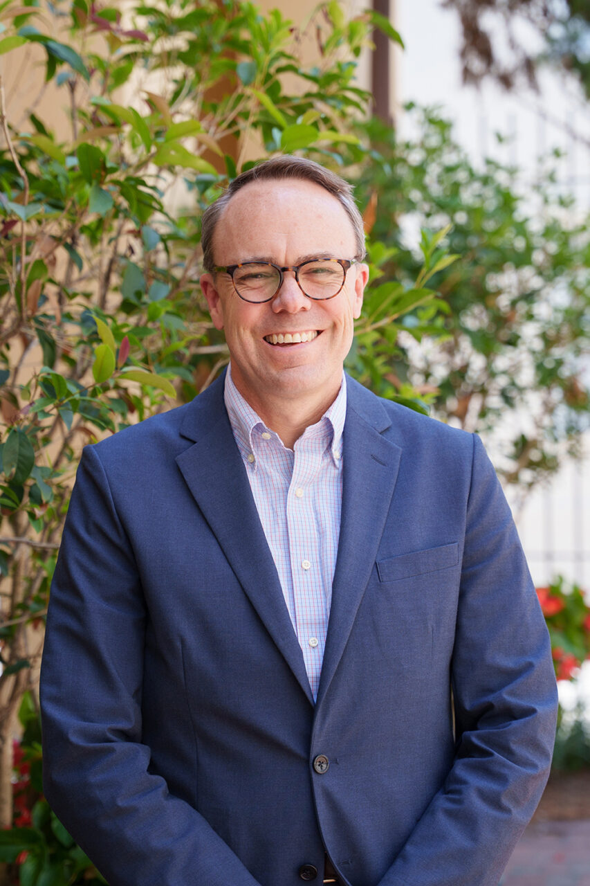 A person in a blue blazer and glasses smiles while standing outdoors near greenery and a building.
