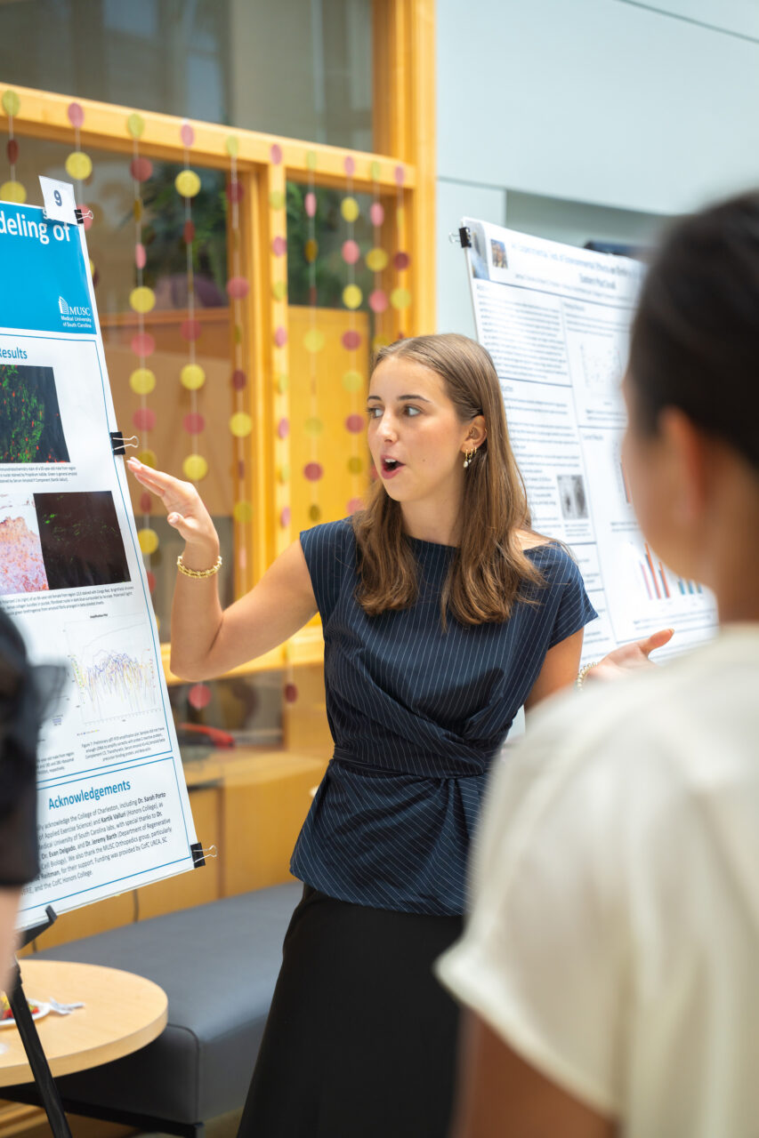 A person presents research findings to an audience at a conference, gesturing toward a poster with scientific images and text.