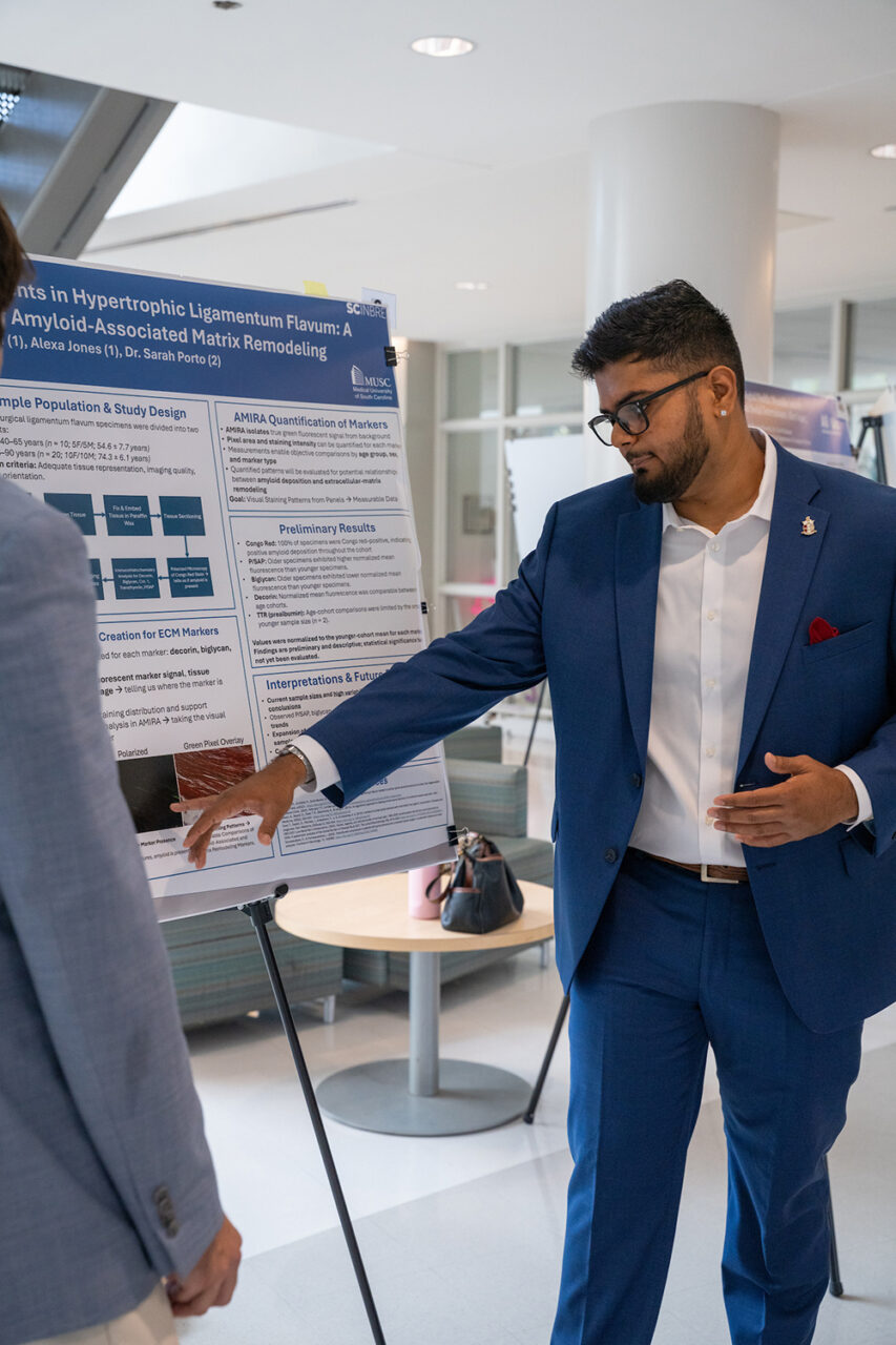 A person in a blue suit gestures toward a scientific research poster while speaking with another individual at what appears to be an academic conference or exhibition.