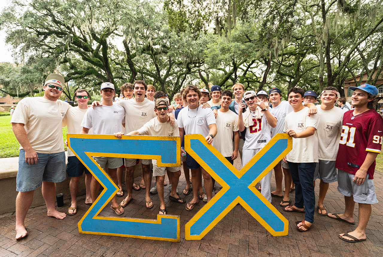 A group of young people pose outdoors with a large blue and yellow Sigma Chi lettering sign.