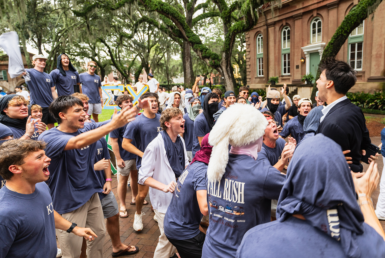 A group of young people in matching blue shirts celebrate energetically outdoors, surrounded by trees and a historic building.