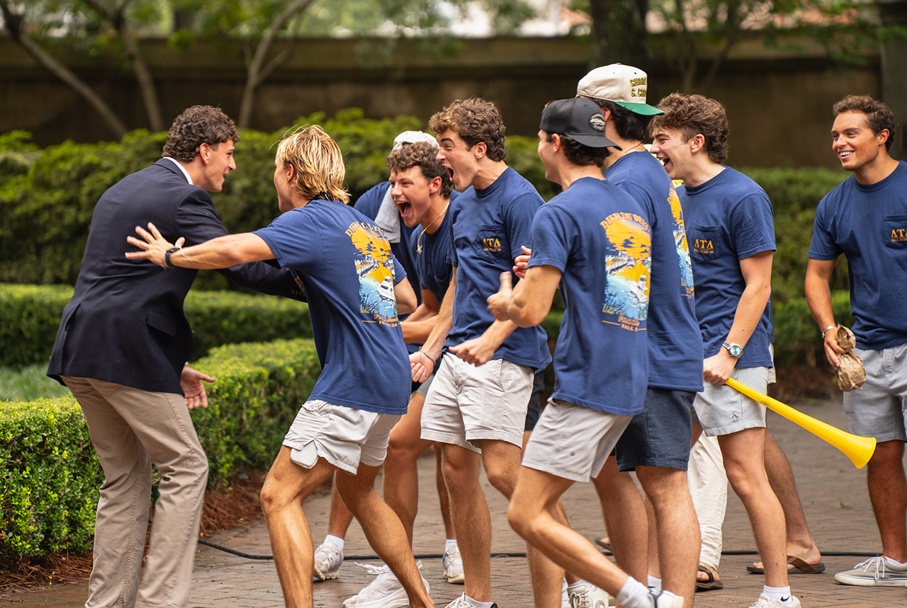 A group of young people in matching blue shirts celebrate outdoors with a man in a suit, one holding a yellow horn.