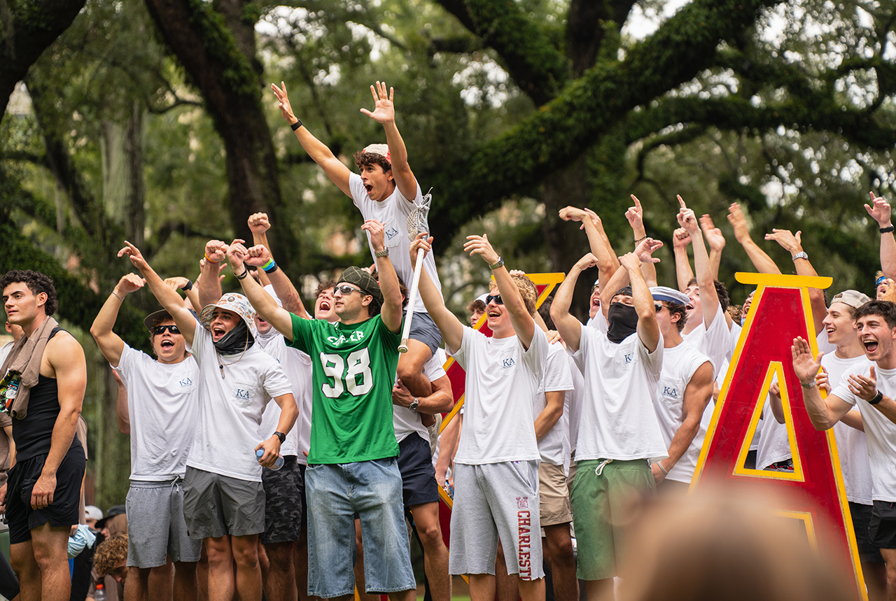 A group of young people celebrate outdoors, raising their arms in excitement near a large red and yellow letter "A".