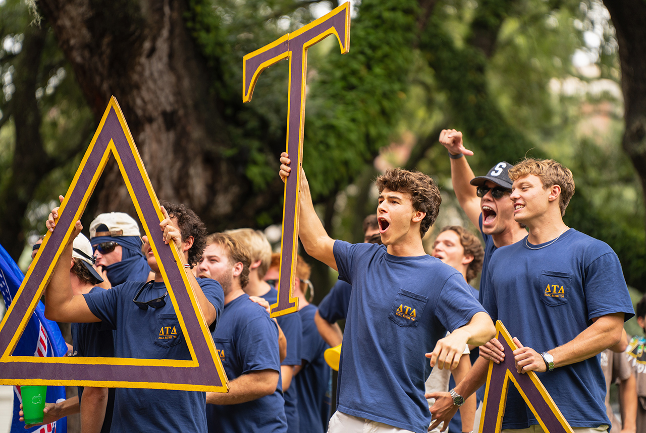 A group of young people in matching blue shirts cheerfully hold large purple and yellow letter signs during an outdoor event.