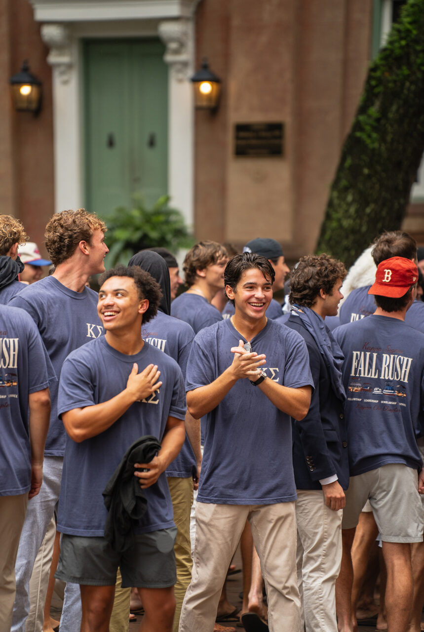 A group of young people in matching blue shirts gather outdoors, smiling and clapping during a Fall Rush event.