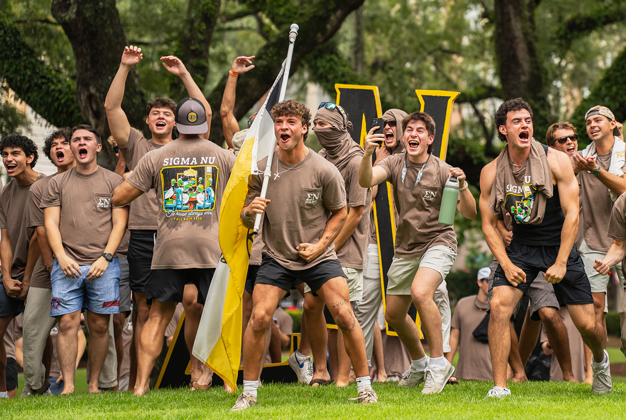 A group of young people in matching brown shirts celebrate enthusiastically outdoors, some holding flags and large letters, with arms raised and joyful expressions.