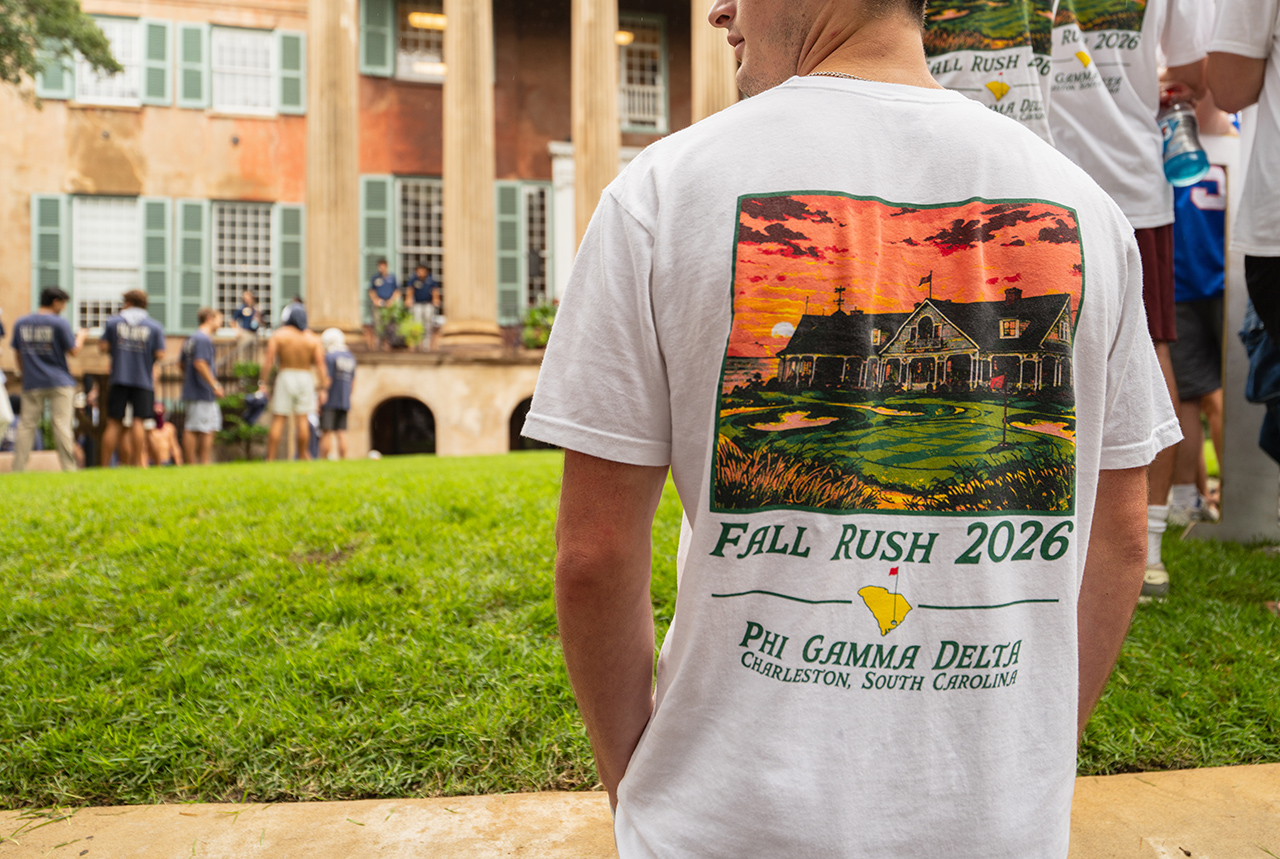 A person wears a white t-shirt featuring a colorful graphic of a house and golf course, with text reading "Fall Rush 2026" and "Phi Gamma Delta, Charleston, South Carolina," standing outdoors near a historic building.