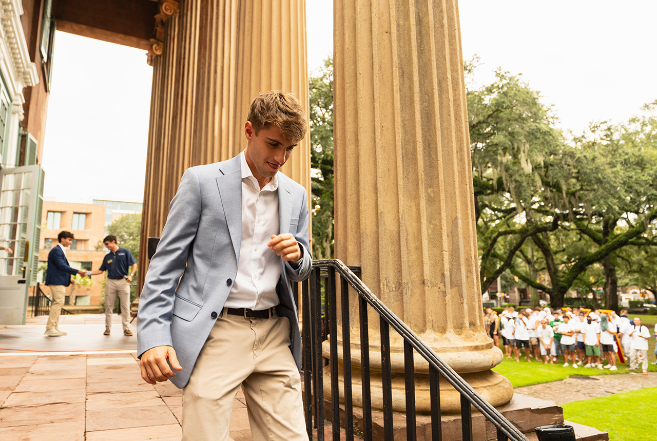 A person in a light blue blazer and khaki pants descends steps beside large columns, with others gathered in the background.